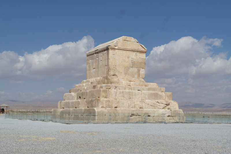 Fotografia przedstawiająca grobowiec Cyrusa Wielkiego w Pasargadach. Na szczycie niewielkiej piramidy schodkowej znajduje się budynek mauzoleum w formie prostego domu z dwuspadowym dachem, zbudowany z kamienia.
